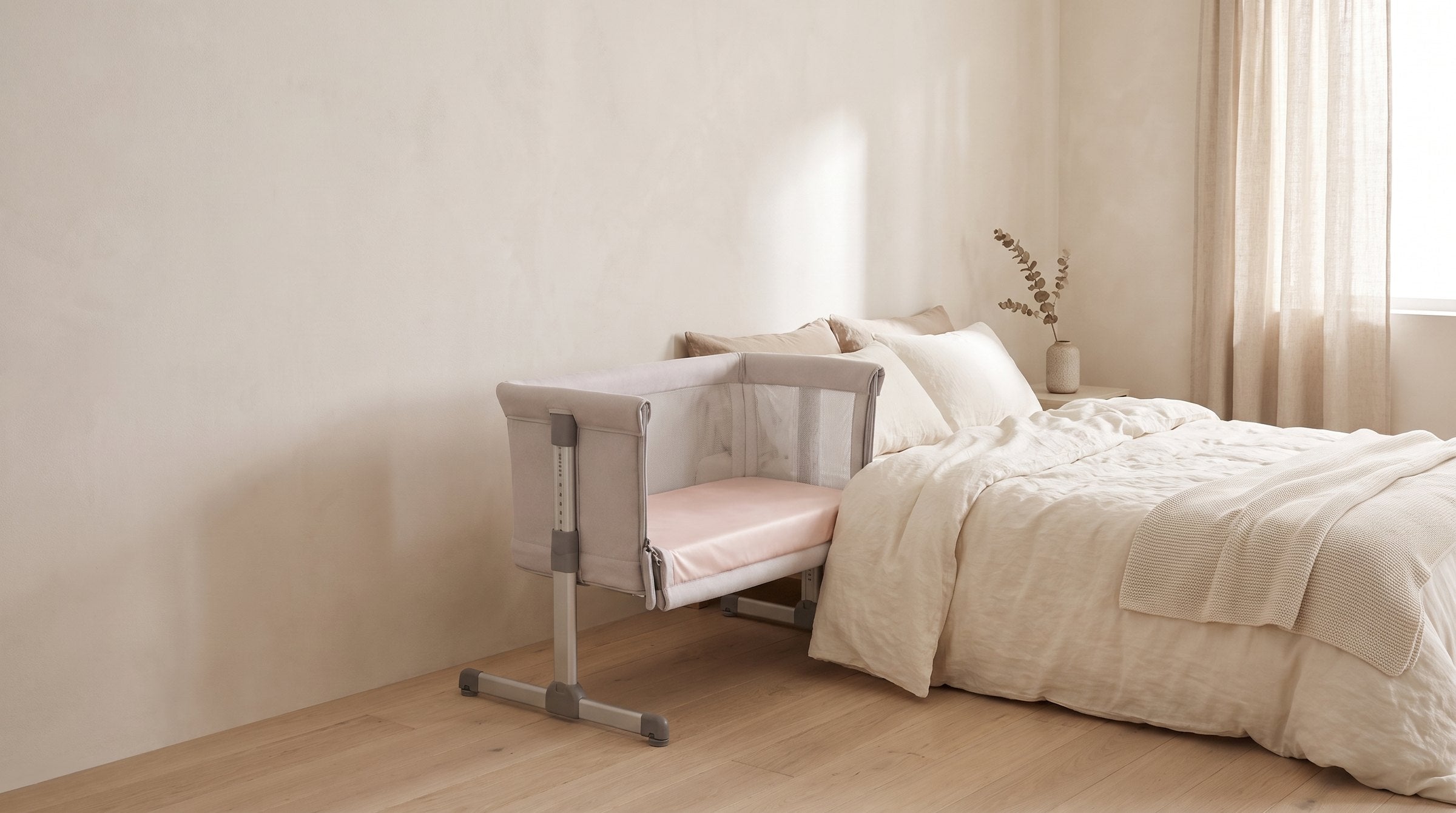 A bedside crib fitted with a Light Pink Sleepy Teddy silk sheet beside a bed in a calm ivory nursery